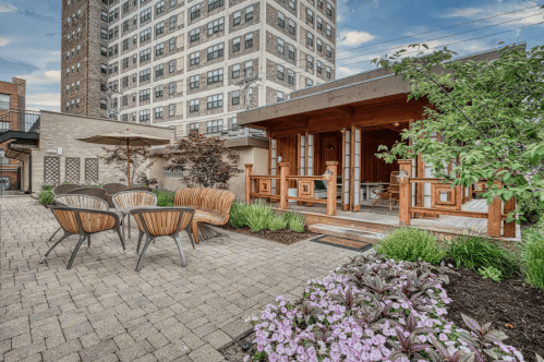 A landscaped outdoor area with wooden seating, a small building, and blooming flowers against a city backdrop.