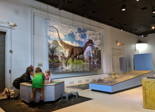 Children interact with a dinosaur exhibit in a museum, featuring a large dinosaur mural in the background.