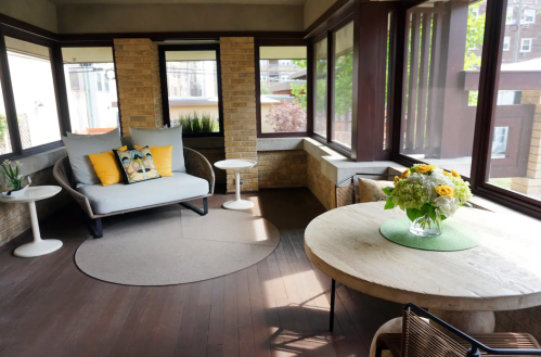 A cozy sunroom with a gray sofa, round table, and floral centerpiece, surrounded by large windows.