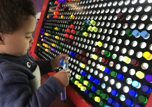 A young child interacts with a colorful light board, inserting tubes into holes of various colors.