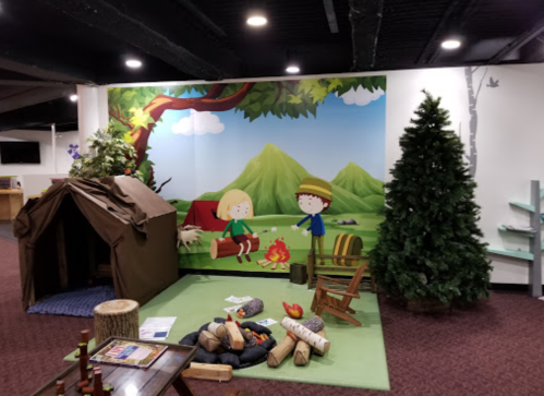 A colorful children's play area featuring a tent, campfire, and a mural of kids in a forest setting.