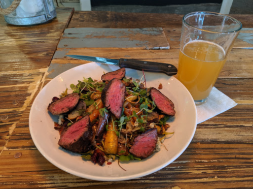 A plate of sliced steak on a bed of greens with vegetables, accompanied by a glass of beer on a wooden table.