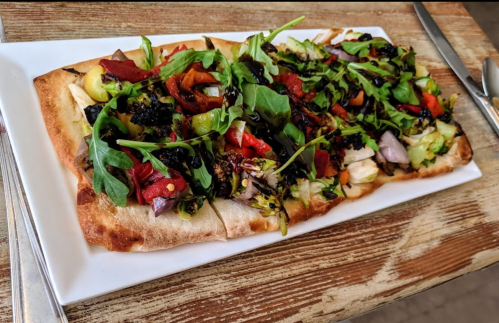A flatbread topped with a colorful mix of greens, roasted vegetables, and herbs, served on a white plate.