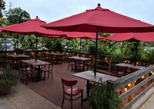 Outdoor dining area with red umbrellas, wooden tables, and chairs surrounded by greenery and soft lighting.