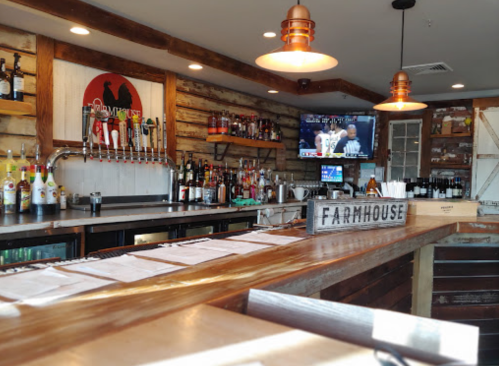 A rustic bar with a wooden counter, shelves of liquor, and a TV showing a sports game in a cozy setting.