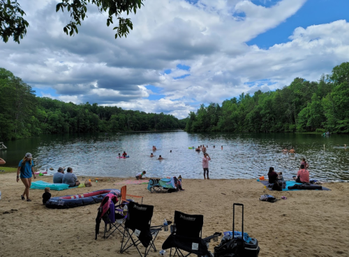A sandy beach by a lake with people swimming and relaxing on the shore under a cloudy sky.