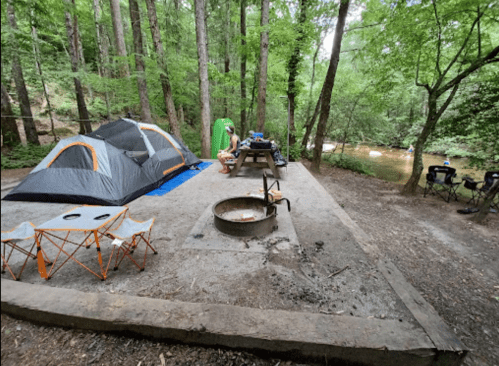A campsite in a wooded area featuring a tent, picnic table, fire pit, and chairs near a river.