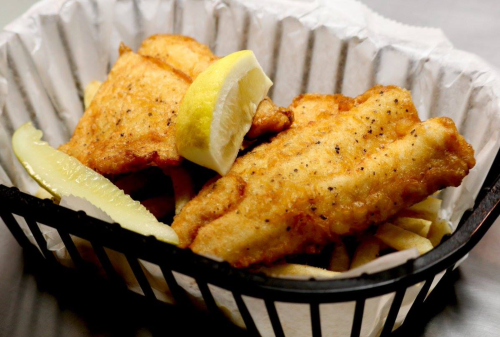 Two pieces of golden fried fish with a lemon wedge, served with fries in a black basket.