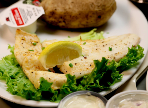 Grilled fish fillets on a bed of lettuce, garnished with a lemon wedge, alongside a baked potato and dipping sauces.