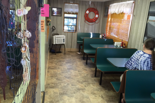 Interior of a casual diner with green booths, a window, and a woman sitting alone. Decor includes seashells and fishing themes.