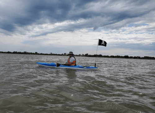 A person in a kayak with a pirate flag paddles on a calm body of water under a cloudy sky.