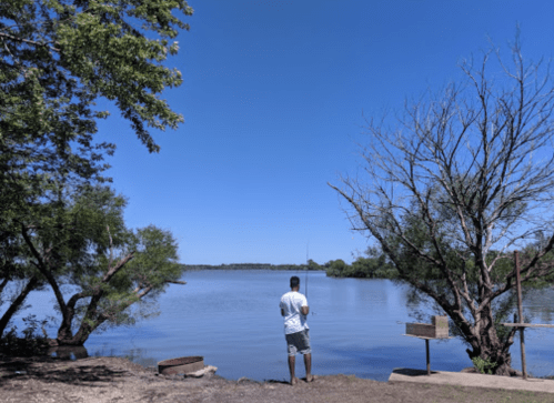 A person fishing by a calm lake under a clear blue sky, surrounded by trees and a peaceful landscape.