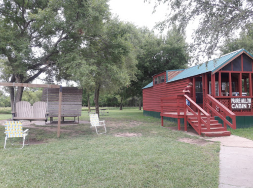 A red cabin with a green roof, surrounded by trees and lawn chairs, with a swing set nearby.