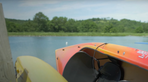 A close-up of a colorful kayak by a calm lake, surrounded by greenery and distant hills.