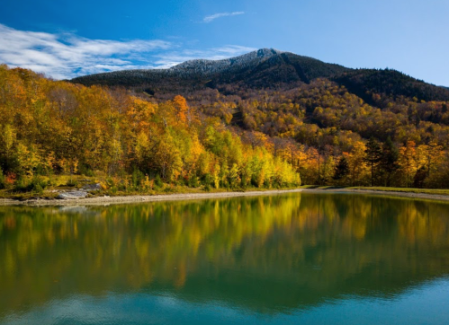 A serene lake reflects vibrant autumn foliage and a mountain under a clear blue sky.