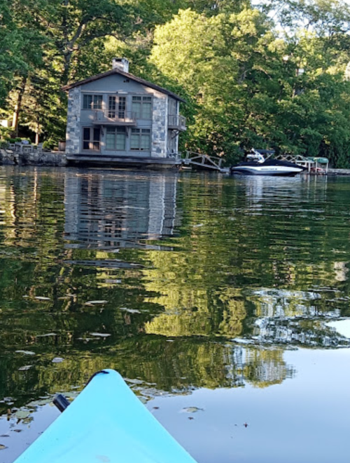A kayak view of a serene lake with a stone house and a boat docked nearby, surrounded by lush green trees.