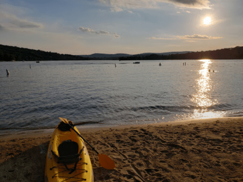 A yellow kayak on a sandy beach by a calm lake at sunset, with distant boats and hills in the background.