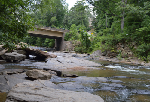 A serene river flows over rocks beneath a bridge, surrounded by lush greenery and trees.