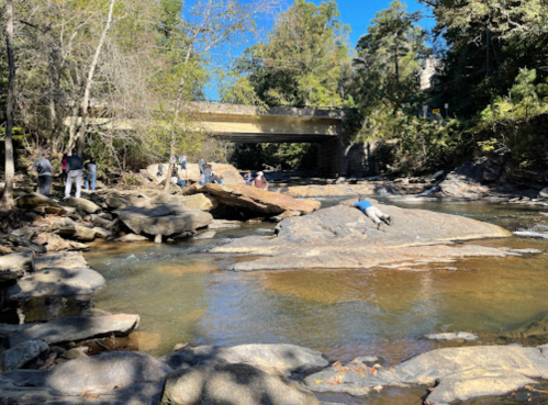 A serene river scene with people exploring rocky banks under a clear blue sky and a bridge in the background.