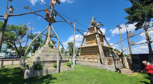 Two large wooden structures with sculptures, surrounded by grass and trees, under a bright blue sky with clouds.