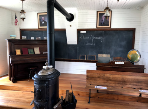 Historic classroom with a wood stove, chalkboard, globe, and wooden benches, featuring portraits on the walls.