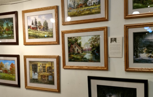 A gallery wall displaying various framed landscape paintings and a photo of an artist with a brief description.