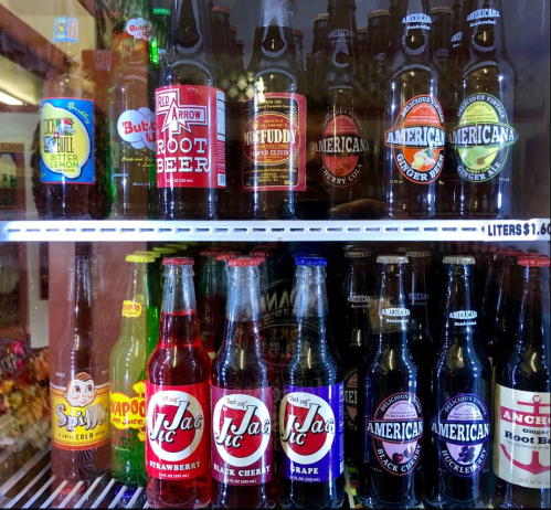 A variety of colorful soda bottles displayed in a glass refrigerator, including root beer, cherry, and grape flavors.