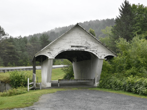 A weathered covered bridge stands in a rural area, surrounded by trees and a misty landscape.