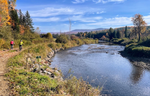 Two cyclists ride along a scenic riverbank surrounded by autumn foliage under a clear blue sky.