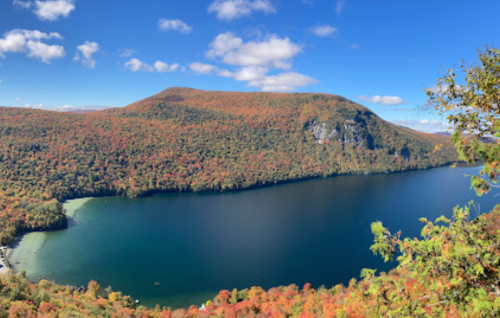A panoramic view of a lake surrounded by vibrant autumn foliage and rolling hills under a blue sky with fluffy clouds.