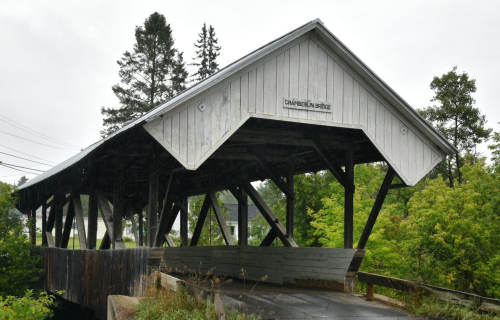 A white, wooden covered bridge with a peaked roof, surrounded by green trees and a cloudy sky.