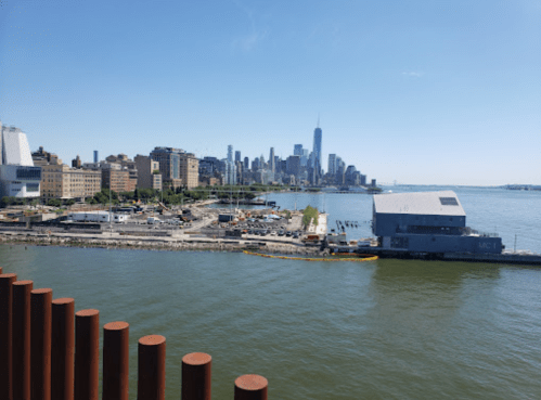 View of a waterfront city skyline with tall buildings and a clear blue sky, featuring a calm river in the foreground.