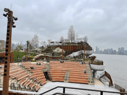 A scenic waterfront park with tiered seating, greenery, and a city skyline in the background under a cloudy sky.