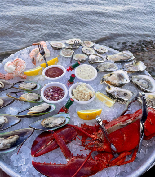 A seafood platter featuring lobster, shrimp, oysters, and mussels on ice, with dipping sauces and lemon wedges.