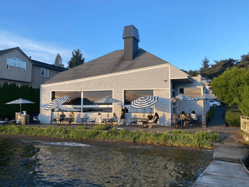 A lakeside restaurant with striped umbrellas, people dining outside, and a clear blue sky in the background.