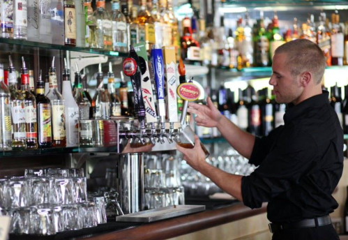 A bartender in a black shirt pours a drink from a tap at a well-stocked bar with various bottles in the background.