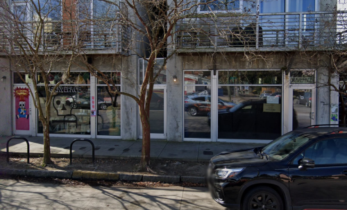 A street view of a storefront with large windows, trees in front, and a parked black vehicle nearby.