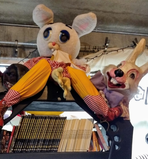 A whimsical display featuring a large stuffed rabbit and a clown-like figure sitting on a shelf with books.