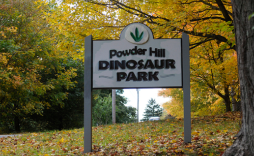 Sign for Powder Hill Dinosaur Park surrounded by colorful autumn foliage.