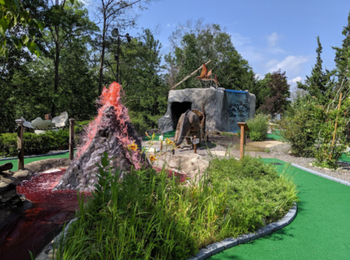 A mini-golf course featuring a volcano, lush greenery, and a cave structure under a clear blue sky.
