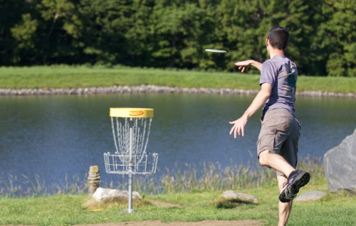 A person throws a frisbee towards a disc golf basket near a pond, surrounded by greenery.