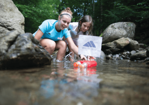 Two girls play in a stream, launching a small boat while surrounded by rocks and greenery.