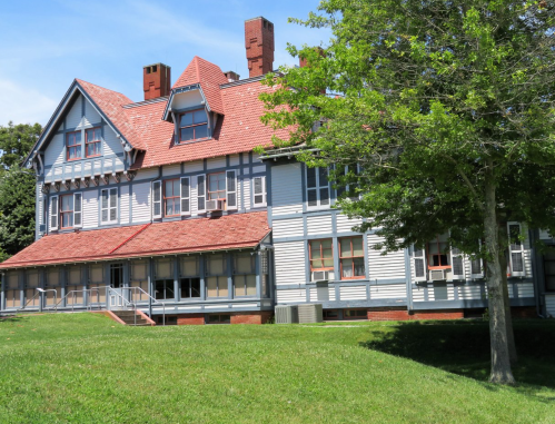 A large, historic building with a red-tiled roof, blue siding, and green lawn, surrounded by trees under a clear sky.