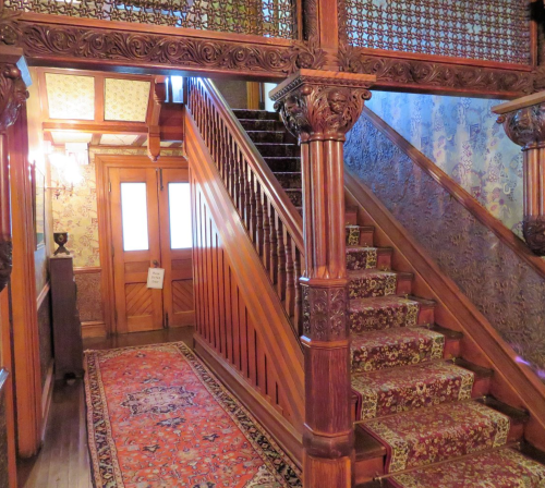 A wooden staircase with ornate carvings, leading to a door, surrounded by patterned wallpaper and a decorative rug.