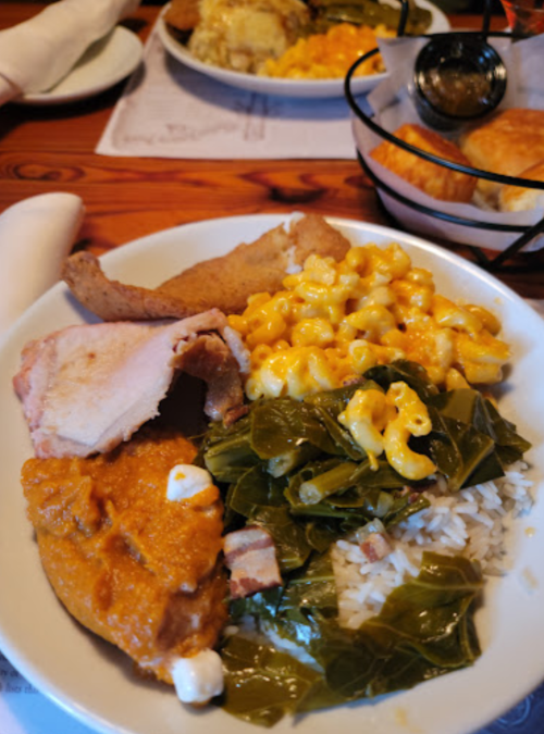 A plate of Southern comfort food featuring fried chicken, collard greens, macaroni and cheese, rice, and sweet potato.