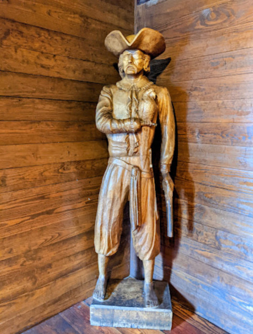 A wooden statue of a pirate stands in a corner, wearing a hat and holding a sword, against a wooden wall.