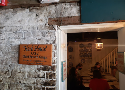 Sign for "Herb House," the oldest house in Georgia, with a glimpse of a dining area and rustic interior.