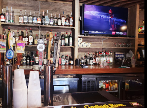 A bar with a variety of liquor bottles on shelves, taps, and a TV displaying a sports event.