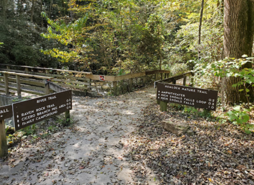 A wooden trail intersection with signs for River Trail, Raven Rock Trail, and Hemlock Nature Trail in a forested area.