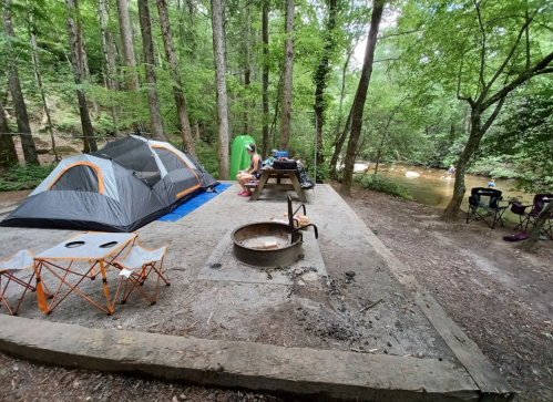A campsite with a tent, picnic table, fire pit, and chairs near a creek, surrounded by lush green trees.